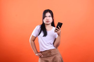 Bir kadın elinde turuncu arka planı olan bir cep telefonu tutuyor ve beyaz gömlek ve bağcıklı kahverengi bir etek giyiyor. Koyu renk saçları var ve yukarı bakıyor.