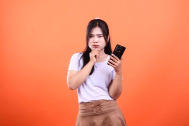 Bir kadın elinde akıllı telefon tutuyor, şaşkın şaşkın bakıyor. Turuncu bir arka planda izole edilmiş..