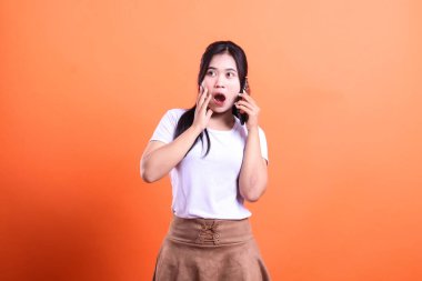 Şaşırmış bir ifade takınan bir kadın elinde bir cep telefonuyla turuncu bir arka plan tutuyor..