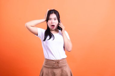 Beyaz gömlekli ve kahverengi etekli bir kadın telefonunu tutuyor ve turuncu bir geçmişi olan bir telefon görüşmesinde şaşırmış görünüyor..