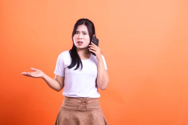 Bir insan elinde bir telefon tutuyor ve kafası karışmış, turuncu bir arkaplanda izole edilmiş bir yüz ifadesi var. Beyaz bir gömlek ve kahverengi bir etek giyiyorlar..