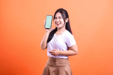 Genç bir kadın gülümsüyor ve elinde mobil uygulamaları ve dijital içeriği gösteren yeşil ekranlı akıllı bir telefon cihazı tutuyor. O, turuncu arka planda izole edilmiş beyaz bir gömlek ve kahverengi bir etek giyiyor..