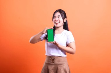 Koyu renk saçlı genç bir kadın yeşil ekranlı bir akıllı telefon tutuyor ve onu turuncu arka planda sunuyor. Beyaz tişört ve kahverengi etek giyiyor, turuncu arka planda izole edilmiş.