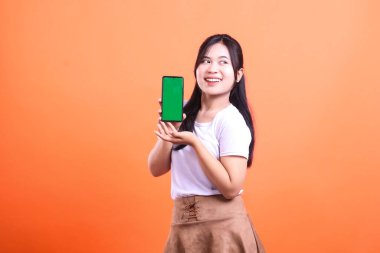 Sağ elinde yeşil ekran olan bir kadın cep telefonunu tutuyor, soluna bakıyor ve gülümsüyor, turuncu arka planda izole bir şekilde..