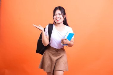 Siyah sırt çantası ve mavi kitabı olan genç bir kadın gülümsüyor ve turuncu arka planda izole edilmiş, avuç içi açık bir şey sunuyor.