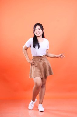 Turuncu bir arka fonda duran bir kadının tam pozu. Beyaz bir tişört, kısa kahverengi bir etek ve beyaz ayakkabılar giyiyor..