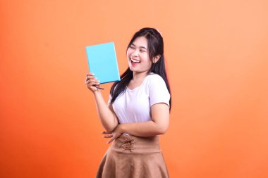 Bir kadın mavi bir kitap tutuyor ve ağzı açık gülümsüyor poz verirken, beyaz bir gömlek ve kahverengi bir etek giyiyor, turuncu arka planda izole edilmiş.