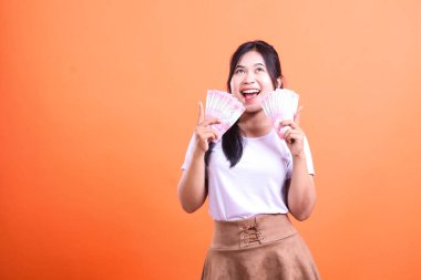 Bir kadın ağzı açık gülümserken iki grup banknot tutuyor. O, turuncu bir arka planın önünde duruyor. O beyaz bir tişört ve kahverengi bir etek giyiyor..