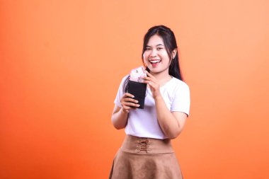 Beyaz tişört ve kahverengi etek giyen gülümseyen bir kadın Endonezya Rupiah banknotlarıyla dolu siyah bir cüzdan tutuyor. turuncu arkaplanda izole