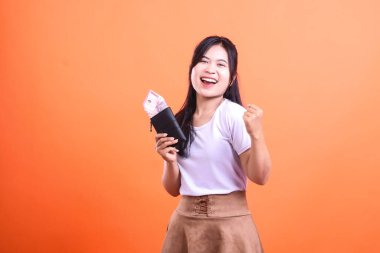 Bir kadın para dolu bir cüzdan tutar ve yumruğunu neşeyle pompalar. O gülümsüyor ve beyaz bir tişört ve kahverengi bir etek giyiyor. Arka plan turuncu..