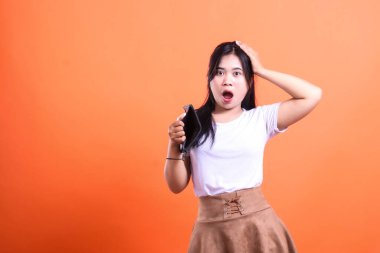 Uzun siyah saçlı ve beyaz tişörtlü bir kadın boş cüzdanını gördüğüne şaşırır. Onun eli kafasında ve ağzı açık. turuncu arkaplanda izole