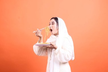 Başörtüsü takan genç bir kadın yemek çubuklarıyla erişte yiyor. Yüzü zevk ve memnuniyet içinde ışıldıyor. turuncu bir arkaplanda izole.
