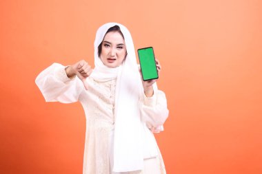 Başörtüsü takan Asyalı bir kadın yeşil ekranlı bir telefonu gösterirken baş parmağıyla yaptığı jestle onaylamadığını ifade ediyor. turuncu bir arkaplanda izole.
