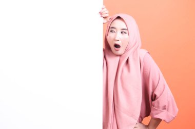 Pembe tesettürlü genç bir Müslüman kadın, beyaz bir panonun arkasına bakıyor. Şaşırmış bir ifadeyle, şokta, turuncu bir arka planda izole bir şekilde..