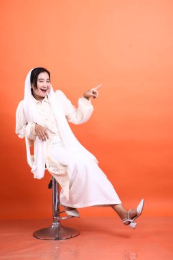 Beyaz tesettürlü neşeli bir Müslüman kadın bir sandalyeye oturur ve yukarı doğru ışıldayan bir gülümsemeyle turuncu bir arka planda izole edilmiş pozitiflik ve yön aktarır..