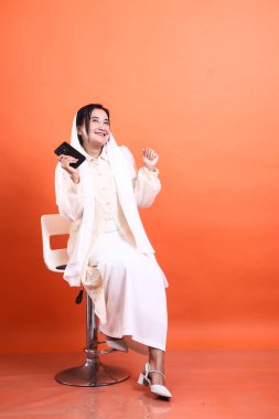 Güzel bir Müslüman kadın tesettürlü gülümsüyor ve akıllı bir telefon tutuyor, pozitiflik ve güven saçıyor, turuncu bir arka planda izole edilmiş, gelenek ve modern teknolojinin bir karışımını sergiliyor..