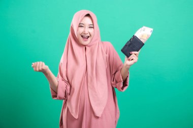 Yeşil arka planda izole edilmiş refah ve finansal başarıyı sembolize eden para dolu bir cüzdan taşıyan tesettürlü neşeli Müslüman kadın. İş başarısını ve kazancını kutluyor..