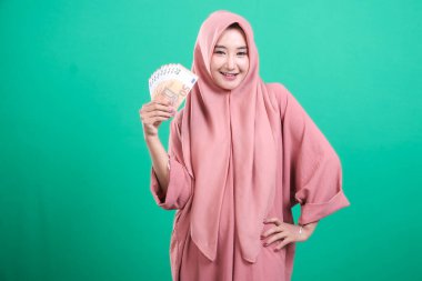 Elinde para tutarken gülümseyen tesettürlü genç Müslüman kadın, yeşil arka planda izole edilmiş girişimlerinde mali güvenliği ve başarıyı simgeliyor. Bu yükselen ekonomiyi vurguluyor.