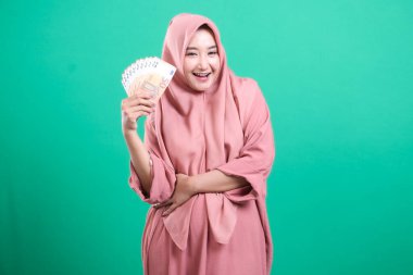 Zengin ve başarıyı zarafetle vurgulayan, avro banknotlarını pembe tesettürle sergileyen neşeli Arap kadın. Yeşil bir zemin üzerinde izole edilmiş, finansal kavramlar ve kültürel temsil için mükemmel, ulaşım.