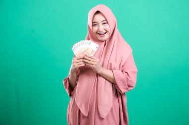 Genç Müslüman kadın, para sergiliyor. Finansal başarının neşeli bir ifadesiyle. Yeşil arka planda izole edilmiş, neşe ve parasal zaferini vurguluyor, başarının bir sembolü..