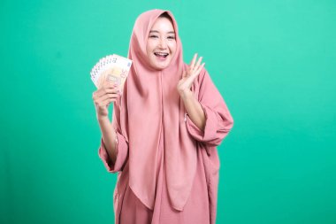 Avro banknotları elinde tutan neşeli Müslüman bir kadın yeşil bir arka planda izole edilmiş canlı bir gülümsemeyle finansal zaferinden duyduğu neşeyi dile getiriyor..