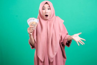 Başörtüsü takan neşeli Müslüman bir kadın, elinde avro banknotları tutuyor, şaşırmış ve yeşil arka planda izole görünüyor. Kazandığı parayla heyecanı ve mutluluğu ifade ediyor..