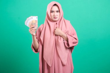 Başörtüsü takan Asyalı bir kadın, yeşil arka planda izole edilmiş, şüpheci bir ifadeyle bir avro hayranı sergiliyor. Ekonomik güvensizlik ve para kaygılarını gösteriyor.