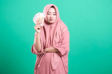 Pembe tesettürlü düşünceli genç müslüman kadın, yeşil arka planda izole edilmiş mali zorluklar göz önüne alındığında, ellerindeki avro banknotlarına üzülerek bakıyor..