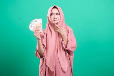 Başörtüsü takan genç bir kadın, yeşil bir arka planda izole edilmiş bir şekilde, bir yığın avro banknotuna dikkatlice bakıyor. Bütçe konusunda endişeli görünüyor..