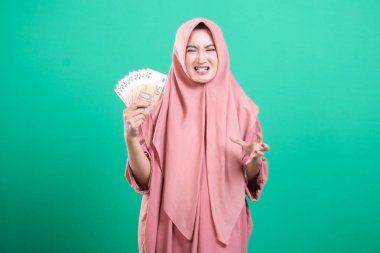 Hiyerarşik giyinmiş hoşnutsuz bir Müslüman kadın nakit tutuyor ve muhtemelen maddi sorunlar nedeniyle hayal kırıklığını dile getiriyor. Yeşil arkaplanda izole edilmiş.