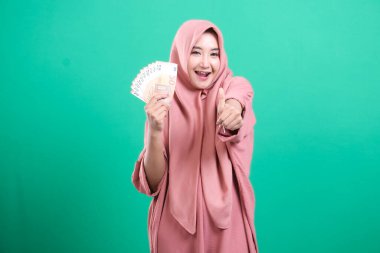 Başparmakları yukarıda, avro parası içinde tesettürlü gülümseyen Müslüman kadın, yeşil arka planda izole edilmiş mali başarı ve başarıyı gösteriyor..