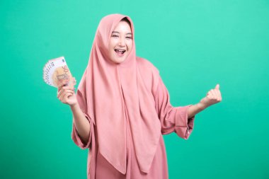 Tesettürlü neşeli Müslüman bir kadın bir stüdyo çekiminde parlak bir şekilde gülümseyen avro para birimi banknotları tutuyor. Yeşil bir arka planda izole edilmiş. Bu görüntü finansal başarının ve refahın özünü yansıtıyor..