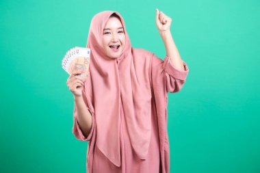 Avro banknotları tutan tesettürlü heyecanlı bir Müslüman kadın yeşil bir zeminde başarı ve mali ödülü veya kazanmayı kutluyor. Kollarını kaldırıp seviniyor. Zaferi ya da başarıyı simgeliyor..