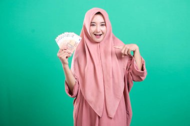 Avro banknotları sunan gülümseyen Müslüman kadın tesettür takıyor, servetinden çok memnun, yeşil bir arka planda izole edilmiş. Zenginlik, başarı ve finansal bağımsızlık kavramı.