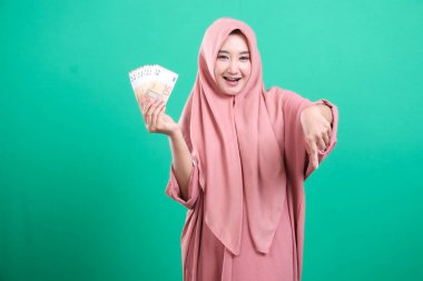 Mutlu Müslüman kadın, yeşil bir arka planda izole edilmiş, sevinçle el kol hareketi yaparken, başarı ve refah gösterisi yaparken avro banknotlarının hayranını tutuyor..