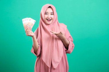 Tesettürlü gülümseyen bir kadın, yeşil arka planda izole edilmiş mali güvenlik ve başarısını ifade ederek avro gösteriyor. Refah, yatırım fırsatları ve parasal refahı temsil eder.