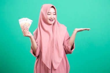 Tesettürlü mutlu Müslüman kadın elinde avro banknotlarıyla gülen bir yüz ifadesiyle refah ve finansal başarıyı temsil ediyor. Yeşil arkaplanda izole edilmiş.