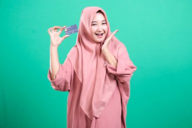 Çevrimiçi alışveriş için kredi kartı tutan tesettürlü tesettürlü Müslüman kadın, yeşil arka planda izole edilmiş neşeli duygularını ifade ediyor. Finans, dijital çağ ve modern Müslüman yaşam tarzı kavramı.