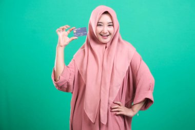 Başörtüsü takan mutlu bir kadın, yeşil arka planda izole edilmiş kolay çevrimiçi ödemeleri ve finansal erişimi simgeleyen bir kredi kartı sergiliyor. Gülüşü, modern bankacılık ve satın alma seçeneklerinin rahatlığını yakalıyor..