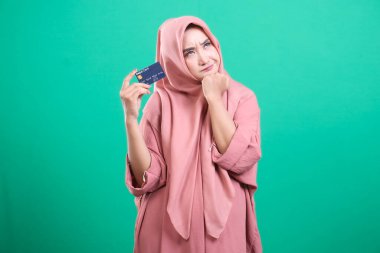 Tesettürlü genç müslüman kadın elinde bir banka kartı tutarken düşünceli görünüyor. Yeşil bir arka planda izole edilmiş harcamalarla ilgili endişelerini dile getiriyor. Finansal zorlukları ve alışverişi yansıtan bir alışveriş yapmayı düşünüyor.