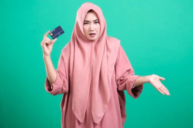 Stresli müslüman kadın yeşil arka planda izole edilmiş, kafası karışık bir ifadeyle kredi kartı gösteriyor. Borç ya da banka hesabı hakkında endişeli bir ifade sergiliyor, şüpheli hareketler yapıyor.