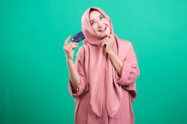 Başörtüsü takan keyifli bir kadın kredi kartı gösteriyor. Çevrimiçi işlemlerin kolaylığını gösteren yeşil bir zeminde izole edilmiş finansal güçlenme ve modern alışveriş alışkanlıklarını öne sürüyor..