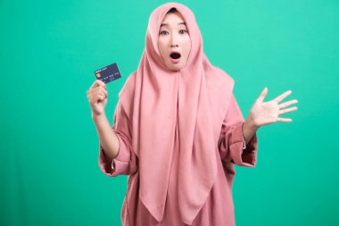 Çevrimiçi harcamalarla ilgili şok olmuş yüz ifadesi ile tesettürlü tesettürlü Müslüman bir kadın, yeşil bir arka planda izole edilmiş. Mali fırsat, kolaylık ve güvenlik sunar..