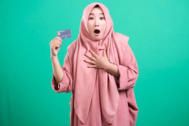 Yeşil bir arka planda izole edilmiş kameraya bakarken elinde kredi kartı olan şaşırmış ve heyecanlı Müslüman bir kadının portresi. Finansal işlemleri karşısında şaşkın ve şaşkın..