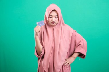 Pembe tesettürlü üzgün ve hayal kırıklığına uğramış Müslüman bir kadın, yeşil bir arka planda izole edilmiş, mali durumu hakkında sıkıntılı bir ifadeyle aşağıya bakan bir kredi kartı tutuyor..