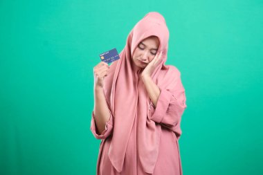 Başörtüsü içinde genç bir Müslüman kadın, elinde kredi kartı tutarken, muhtemelen maddi zorluklar veya borçlarla karşı karşıya, yeşil bir arka planda izole bir şekilde umutsuz görünüyor. Görüntü, ekonomik kaygıları gözler önüne seren anlaşılabilir bir senaryo gösteriyor..
