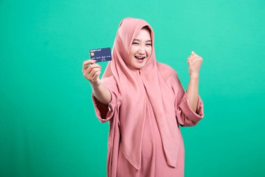 Elinde kredi kartıyla gülümseyen ve neşeli bir Müslüman kadın, başarıyı ve finansal özgürlüğü kutluyor, yeşil bir arka planda izole edilmiş, modern bir yaşam tarzı ve ticaret yoluyla güçlenmeyi sergiliyor.