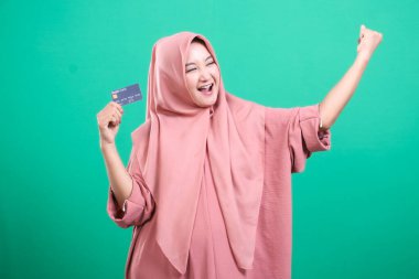 Parasal başarıyı kutlayan neşeli Müslüman kadın, elinde bir kredi kartı tutarken ve kolunu sevinçle kaldırırken, yeşil bir arka planda izole bir şekilde. Mutluluğu, modern bankacılık çözümlerini ve ekonomik olanakları yansıtıyor..