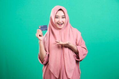 Hevesli kadın tesettürlü bir banka kartı sergiliyor, kusursuz online alışveriş ve ulaşılabilir bankacılık çözümleri sunuyor, yeşil arka planda izole edilmiş, modern bir yaşam tarzı sunuyor..