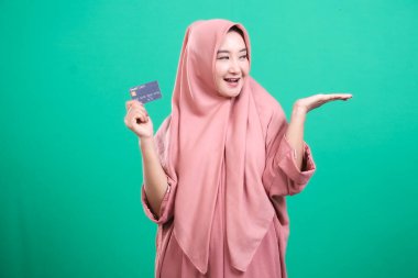 Tesettürlü gülümseyen Müslüman kadın yeşil arka planda izole edilmiş ürün yerleştirme ve promosyon için avuç içi açık bir kredi kartı sunuyor. Finansal okuryazarlığı ve modern kolaylığı temsil eder.